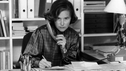 Elisabeth Kopp assise à un bureau, tenant un téléphone fixe d’une main et écrivant de l’autre, entourée de dossiers, de livres et de papiers dans un bureau.