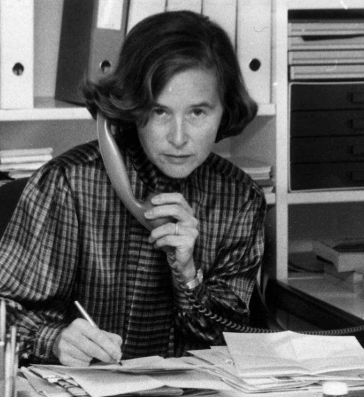 Elisabeth Kopp assise à un bureau, tenant un téléphone fixe d’une main et écrivant de l’autre, entourée de dossiers, de livres et de papiers dans un bureau.
