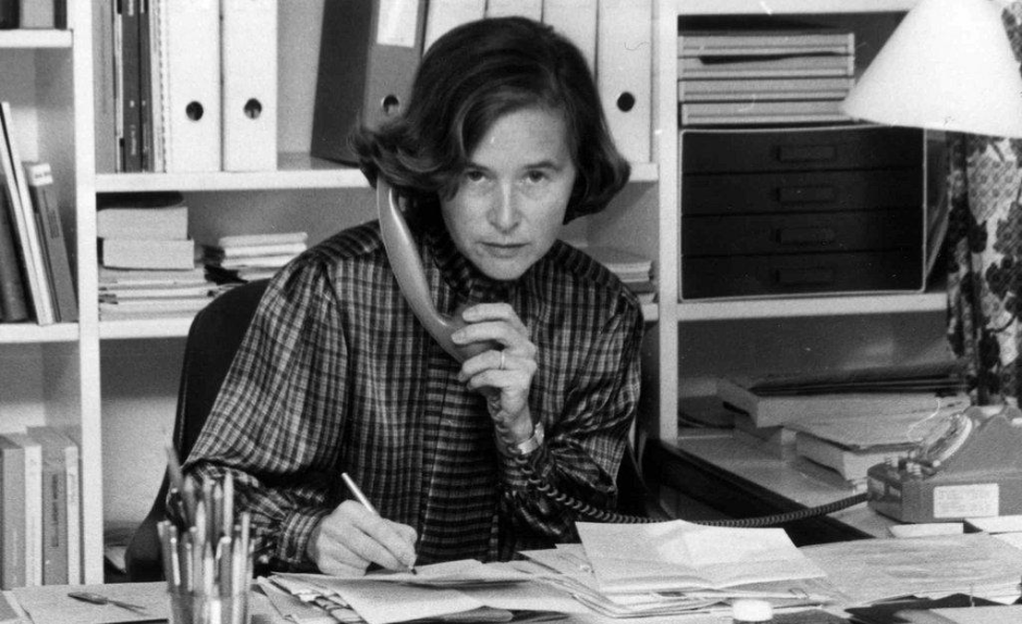 Elisabeth Kopp assise à un bureau, tenant un téléphone fixe d’une main et écrivant de l’autre, entourée de dossiers, de livres et de papiers dans un bureau.