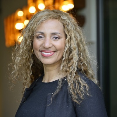 Portrait de Leonilda Renaldo souriante aux cheveux bouclés blonds, portant un haut noir, devant un arrière-plan intérieur flou et chaleureux.