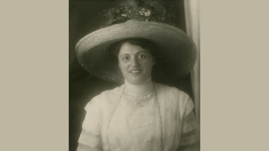 Portrait en noir et blanc de rosa Bloch, femme souriante portant un très large chapeau de paille, vêtue d’une robe claire à col haut, photographiée en buste devant un arrière-plan sombre.
