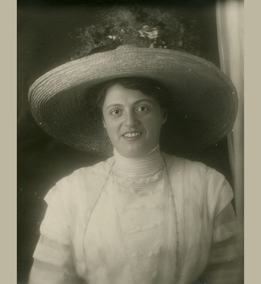 Portrait en noir et blanc de rosa Bloch, femme souriante portant un très large chapeau de paille, vêtue d’une robe claire à col haut, photographiée en buste devant un arrière-plan sombre.