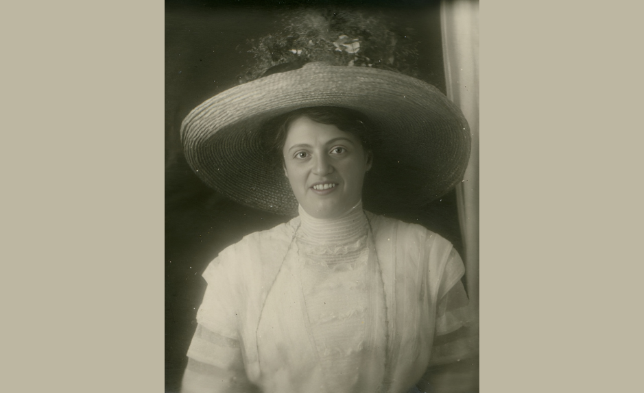 Portrait en noir et blanc de rosa Bloch, femme souriante portant un très large chapeau de paille, vêtue d’une robe claire à col haut, photographiée en buste devant un arrière-plan sombre.