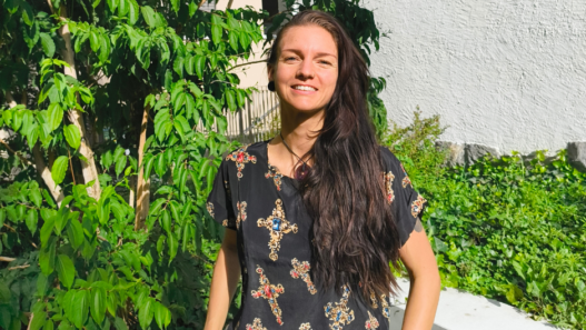 Portrait de Soraya Poulin souriante en extérieur, devant des plantes vertes et un mur blanc.