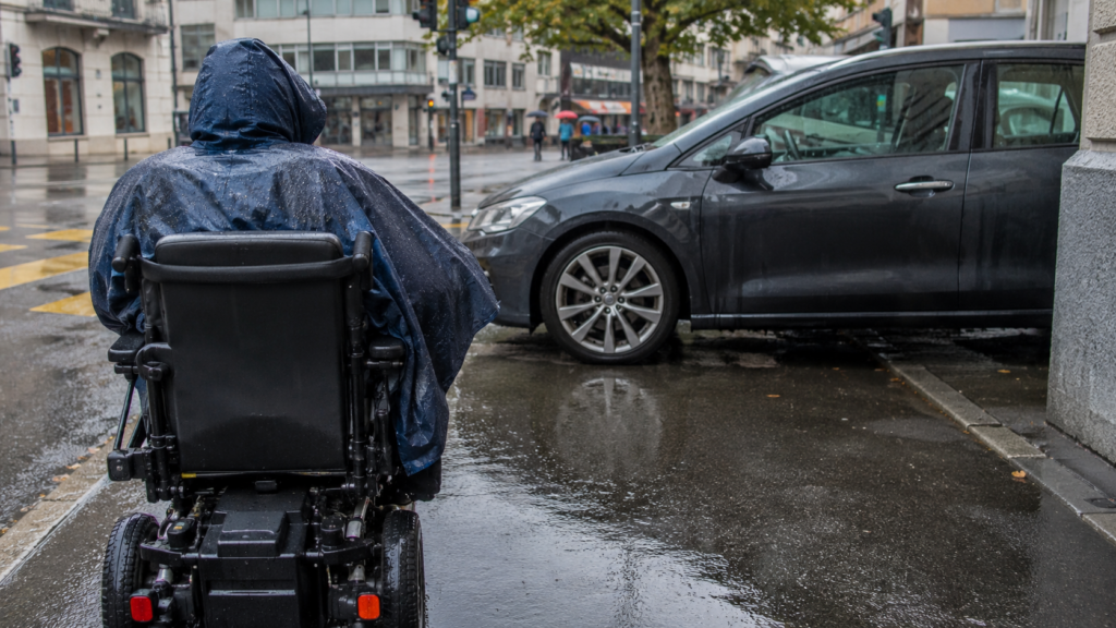 Personne en fauteuil roulant sous la pluie, bloquée par une voiture garée sur le trottoir en ville.