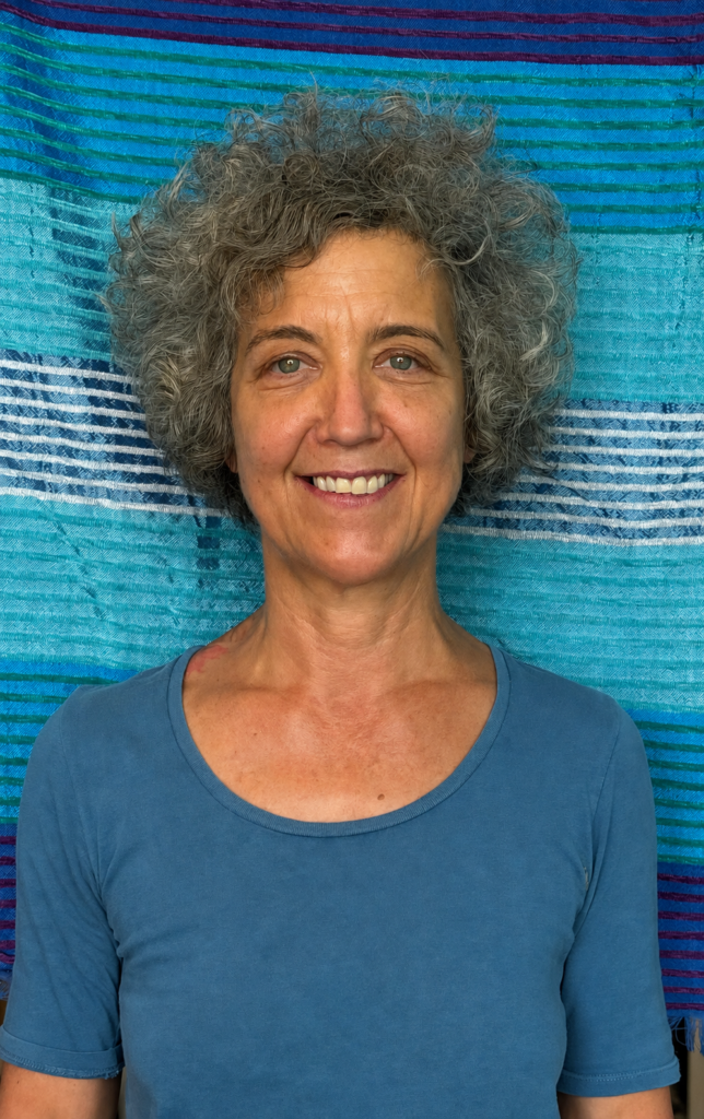 Portrait de Christine Fayet, souriante, aux cheveux gris bouclés, portant un haut bleu devant un fond textile bleu à rayures.
