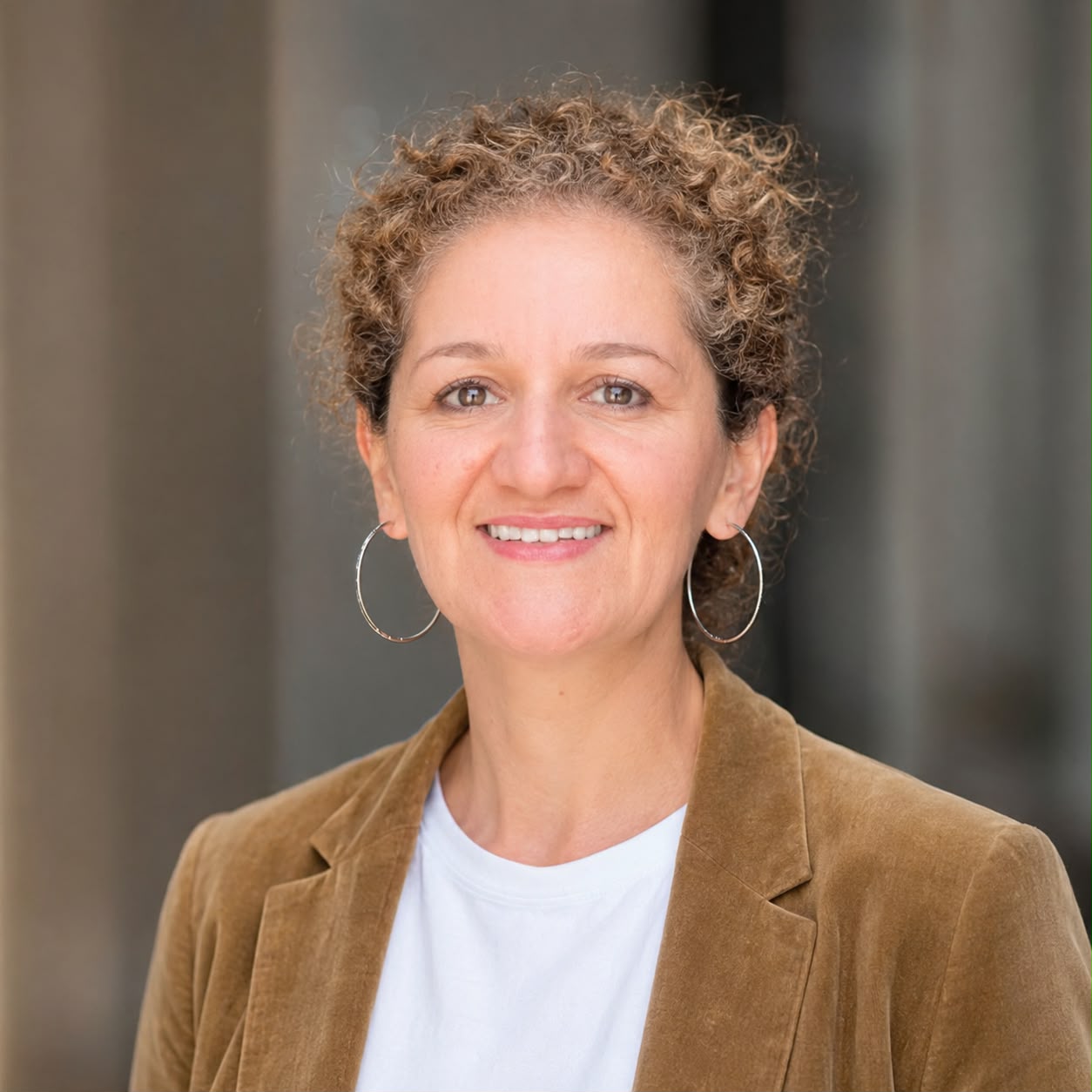 Portrait de Nacèra Mouhoub, réalisatrice et technicienne audiovisuelle, souriante, portant une veste marron sur un haut blanc, devant un arrière-plan urbain flou.