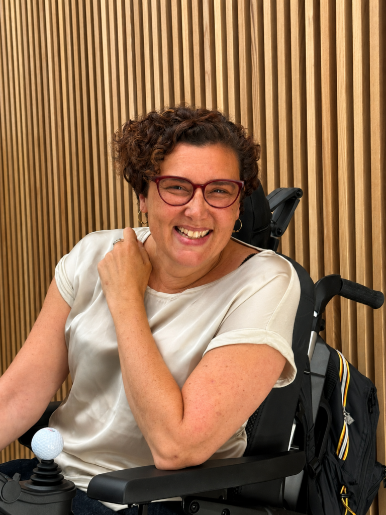 Julie Champenois souriante, assise en fauteuil roulant, devant un fond en bois clair.