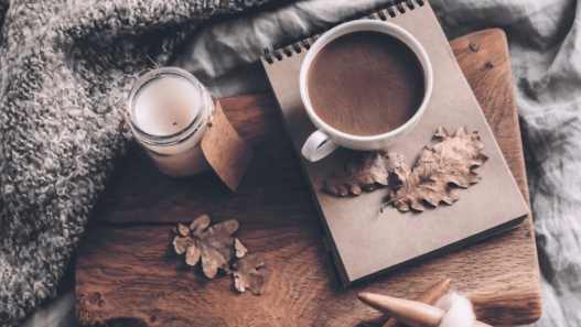 Tasse de café chaud, bougie, carnet et feuilles d’automne près d’un tricot en laine épaisse sur une couverture.