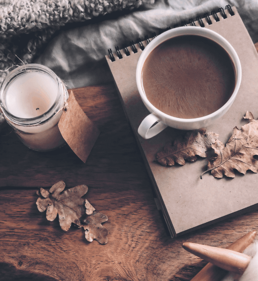 Tasse de café chaud, bougie, carnet et feuilles d’automne près d’un tricot en laine épaisse sur une couverture.