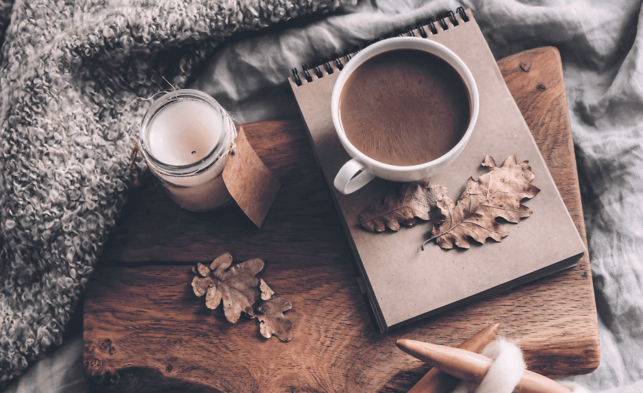 Tasse de café chaud, bougie, carnet et feuilles d’automne près d’un tricot en laine épaisse sur une couverture.