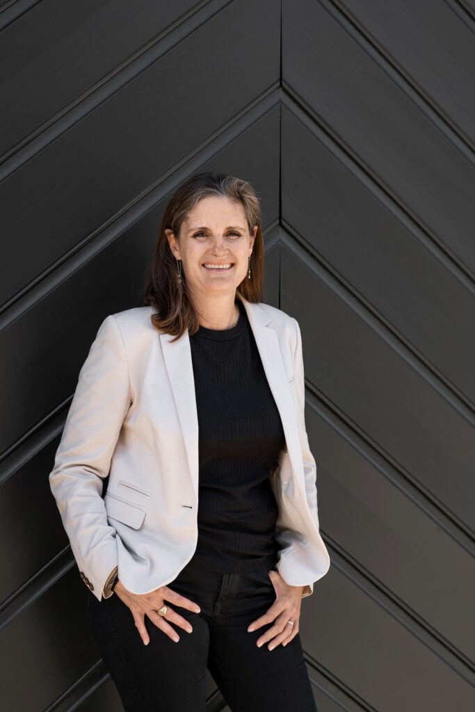 Portrait de Sarah Genequand Miche, gestionnaire de fortune indépendante et autrice, debout devant un mur sombre à motifs géométriques.