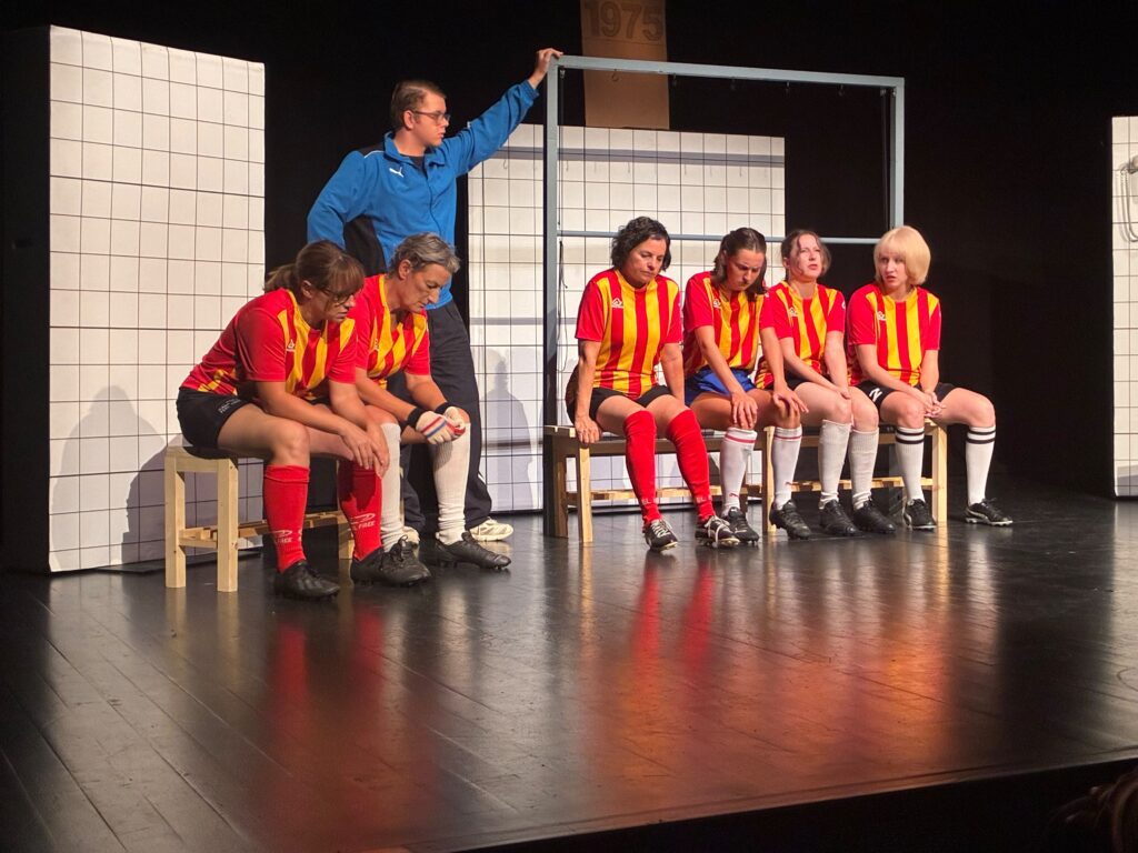 Joueuses en maillots rouges et jaunes assises sur les bancs pendant une scène de Féminines au Théâtre de l’Arbanel à Treyvaux