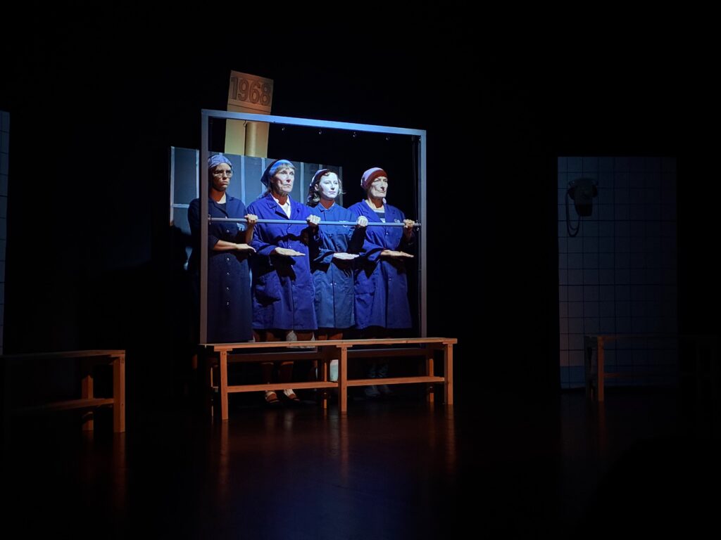 Quatre personnages en blouses bleues sur scène sous l’indication 1968 dans Féminines à Treyvaux