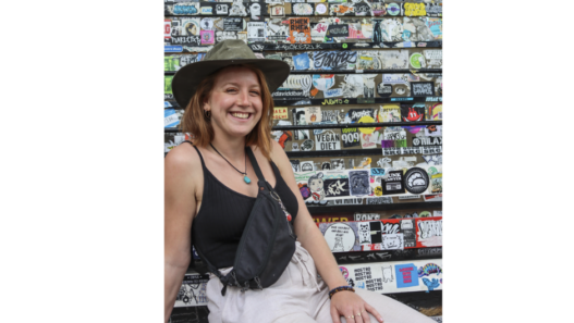 Laure Kohler, autrice jurassienne, souriante et coiffée d’un chapeau kaki, devant un mur couvert d’autocollants et de graffitis.