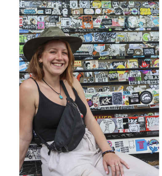 Laure Kohler, autrice jurassienne, souriante et coiffée d’un chapeau kaki, devant un mur couvert d’autocollants et de graffitis.
