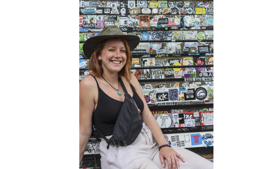Laure Kohler, autrice jurassienne, souriante et coiffée d’un chapeau kaki, devant un mur couvert d’autocollants et de graffitis.